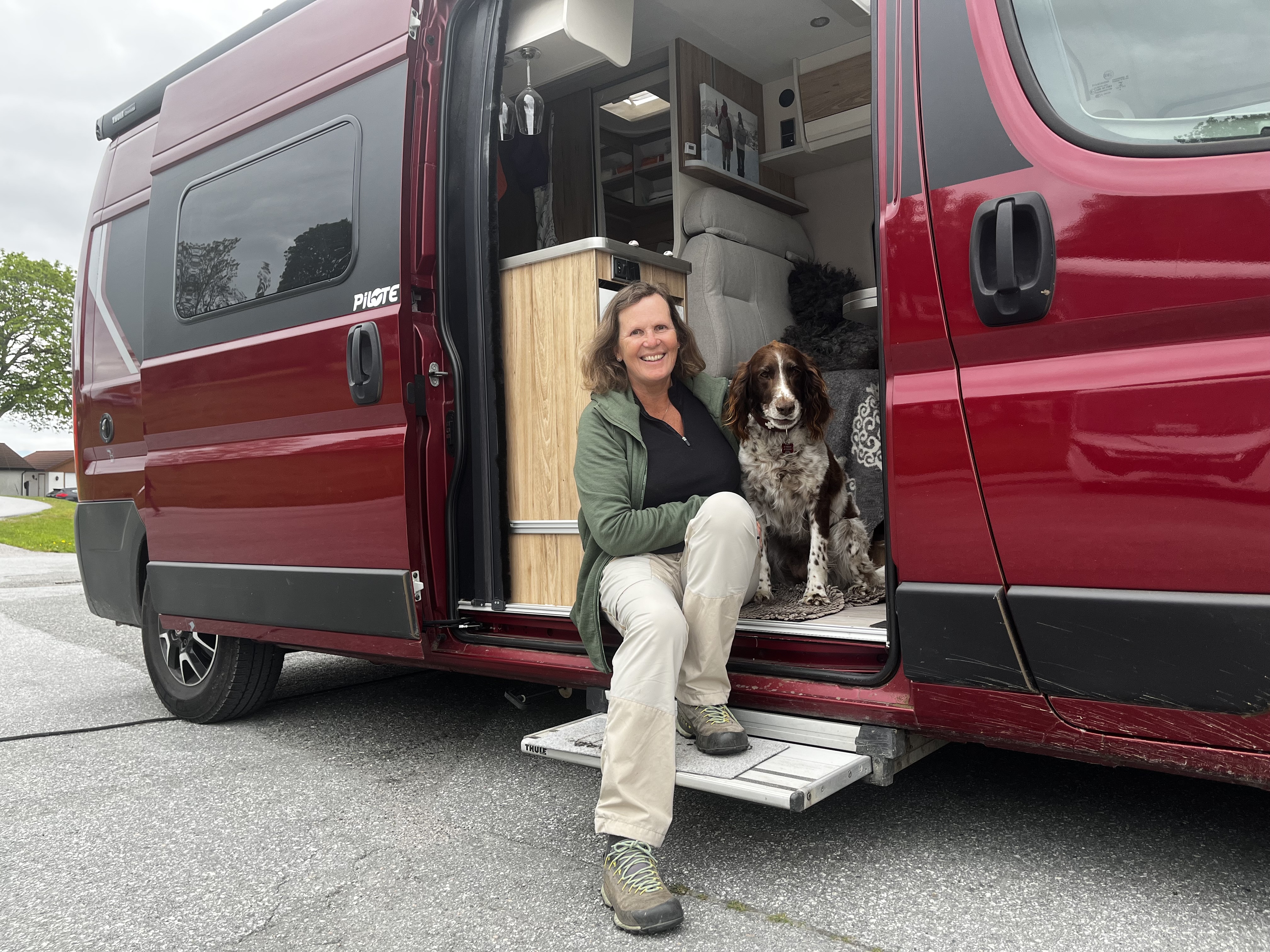 Bildet viser en kvinne og en brun og hvit hund som sitter i sideåpningen på en campervan.